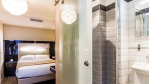 A modern hotel room with a bed, bedside table, and a shower area with a glass door. - District Republique (Paris)