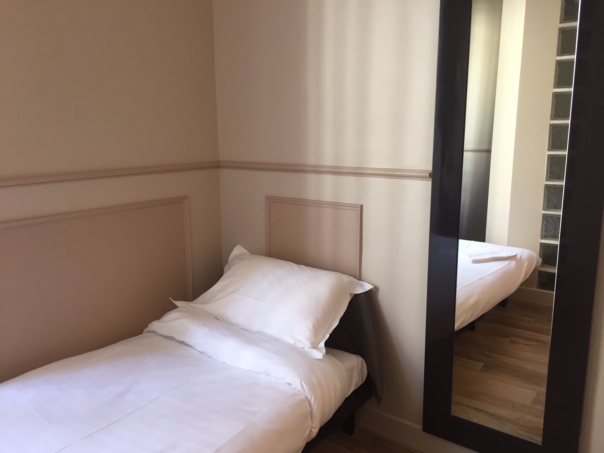 A single bed with white bedding in a room with a wooden floor and a mirror reflecting a staircase.