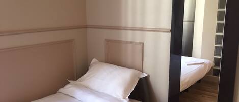 Una cama individual con ropa de cama blanca en una habitación con piso de madera y un espejo que refleja una escalera.