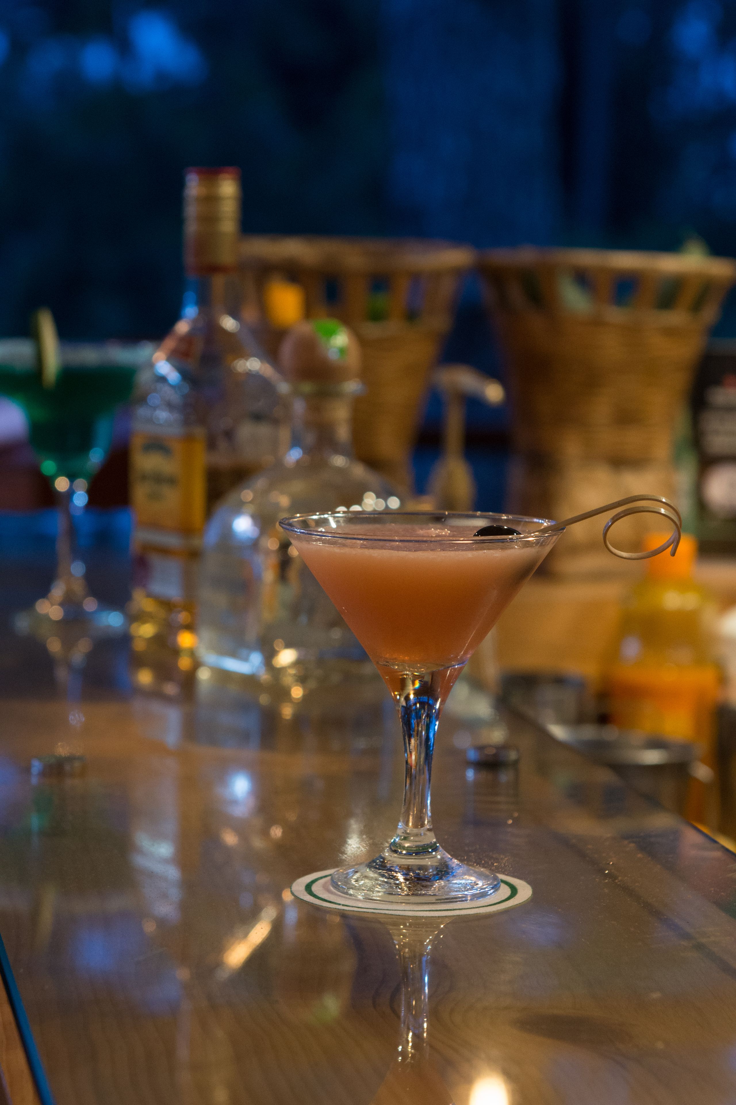 A bar setting with a martini glass filled with a pinkish drink, a bottle of liquor, and a basket in the background.
