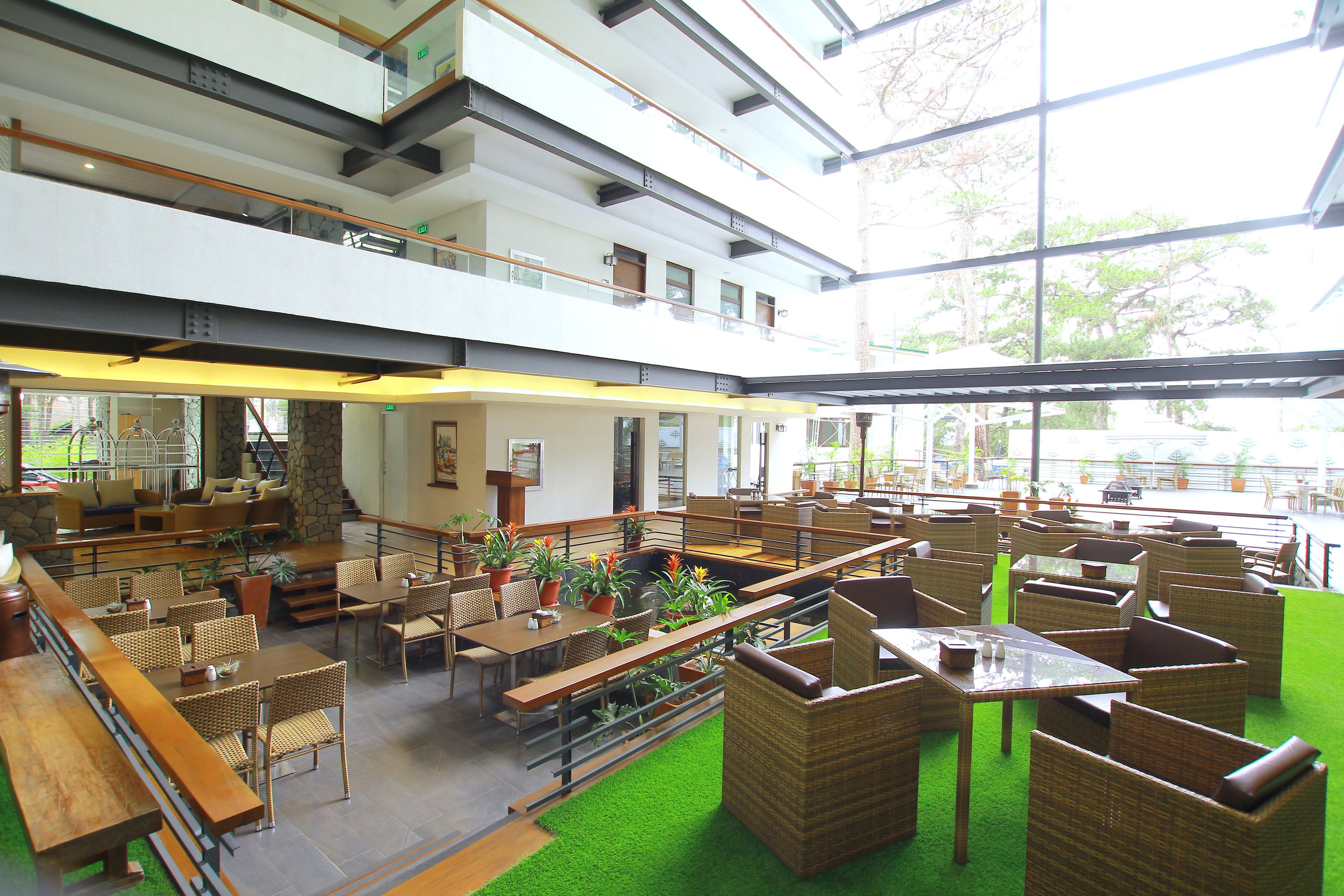 A spacious indoor dining area with multiple tables and chairs, a green carpet, and large windows allowing natural light.
