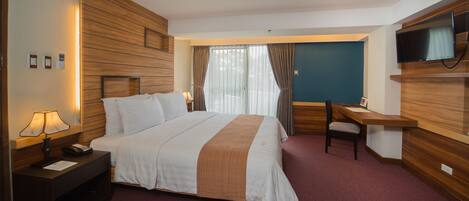Una habitación de hotel con una cama grande, una mesita de noche con lámpara, escritorio, silla y televisor instalado en la pared.