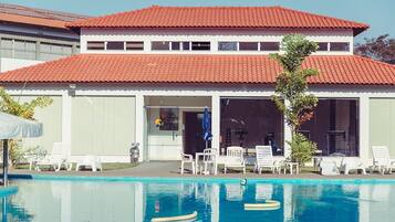 Un bâtiment à deux étages, doté d’un toit recouvert de tuiles rouges, d’une piscine avec des chaises longues et d’un patio couvert.