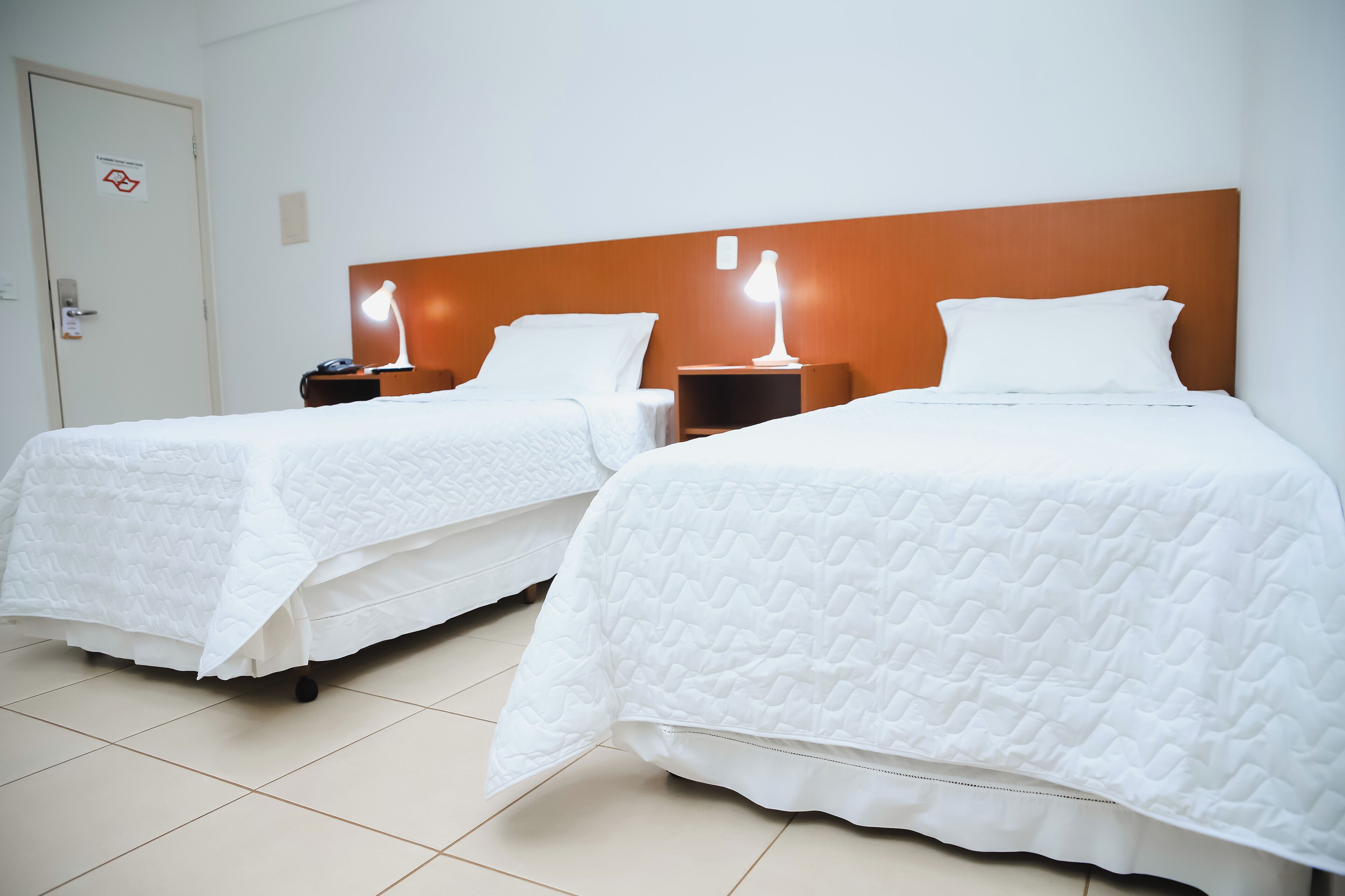 Dos camas individuales con ropa blanca, cabeceros de madera, mesitas de noche con lámparas y piso de baldosas.