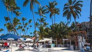 A beach with palm trees, beach chairs, umbrellas, and a small kiosk. - Corales Colonial II With Private Beach,los Corales (Punta Cana)