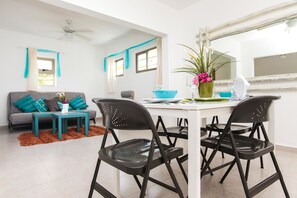 Una sala moderna con un ventilador de techo blanco, cortinas color turquesa, un sofá gris con cojines color turquesa, una mesa de centro blanca y dos sillas negras.