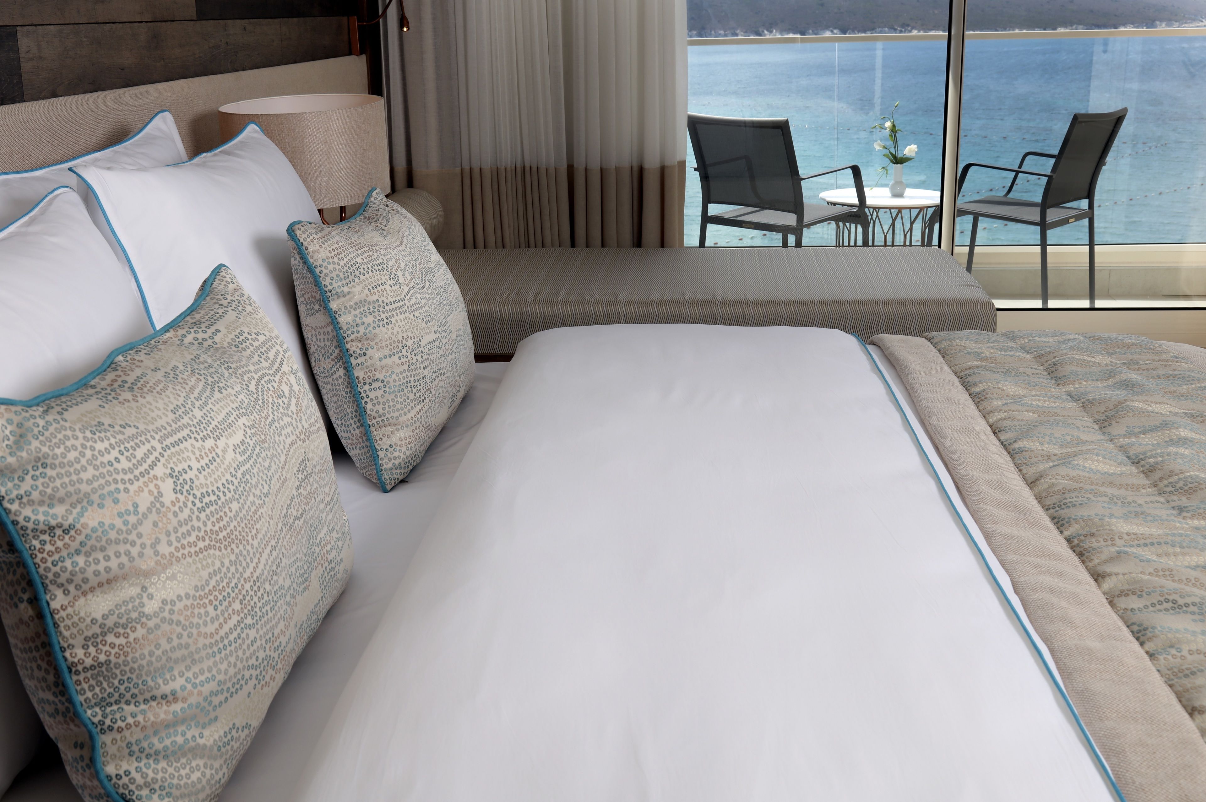 A hotel room with a large bed, two pillows, a bedside table, a lamp, and a view of the sea through large windows.