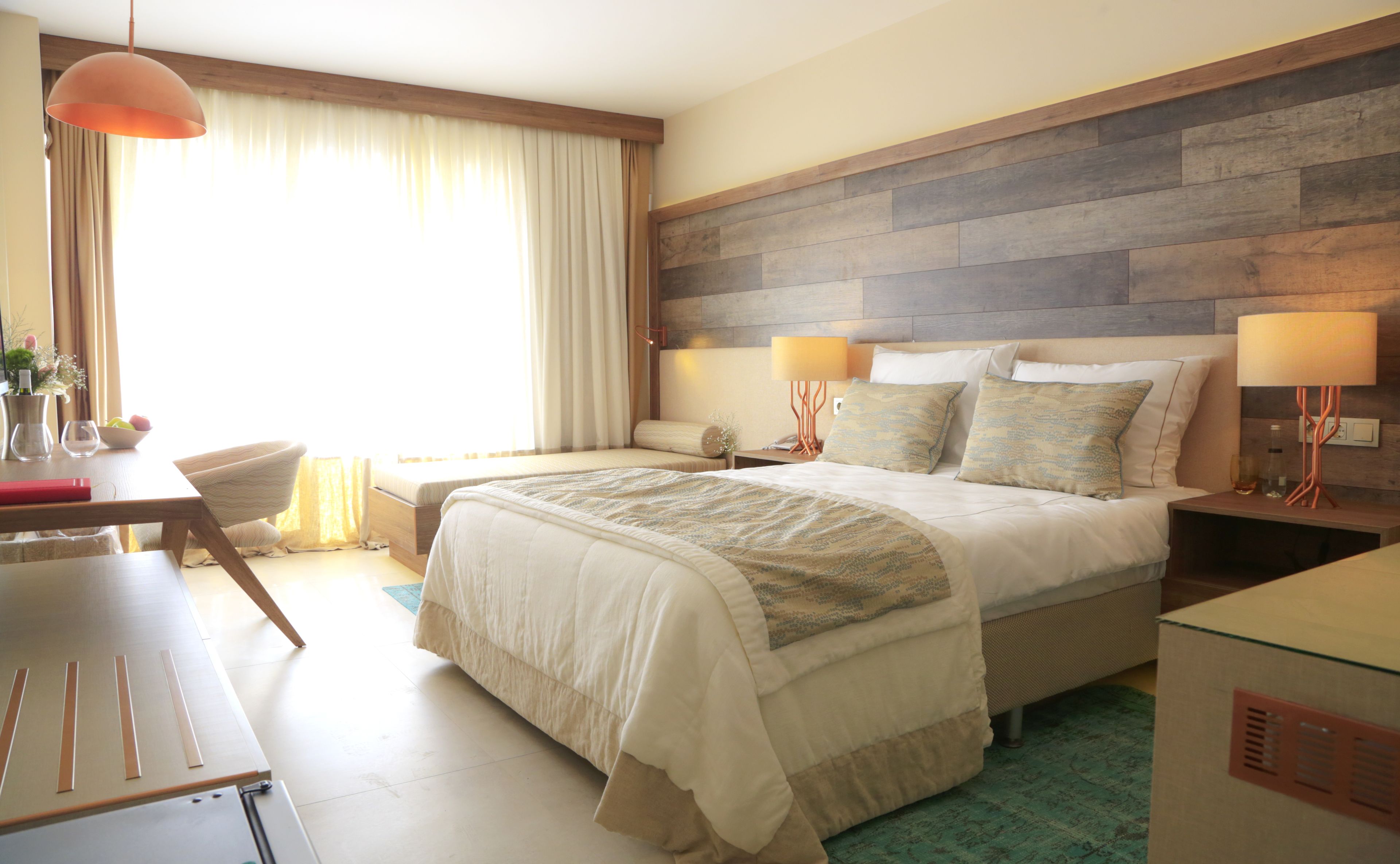 A hotel room with a large bed, bedside lamps, a wooden headboard, a desk with a chair, and a window with curtains.