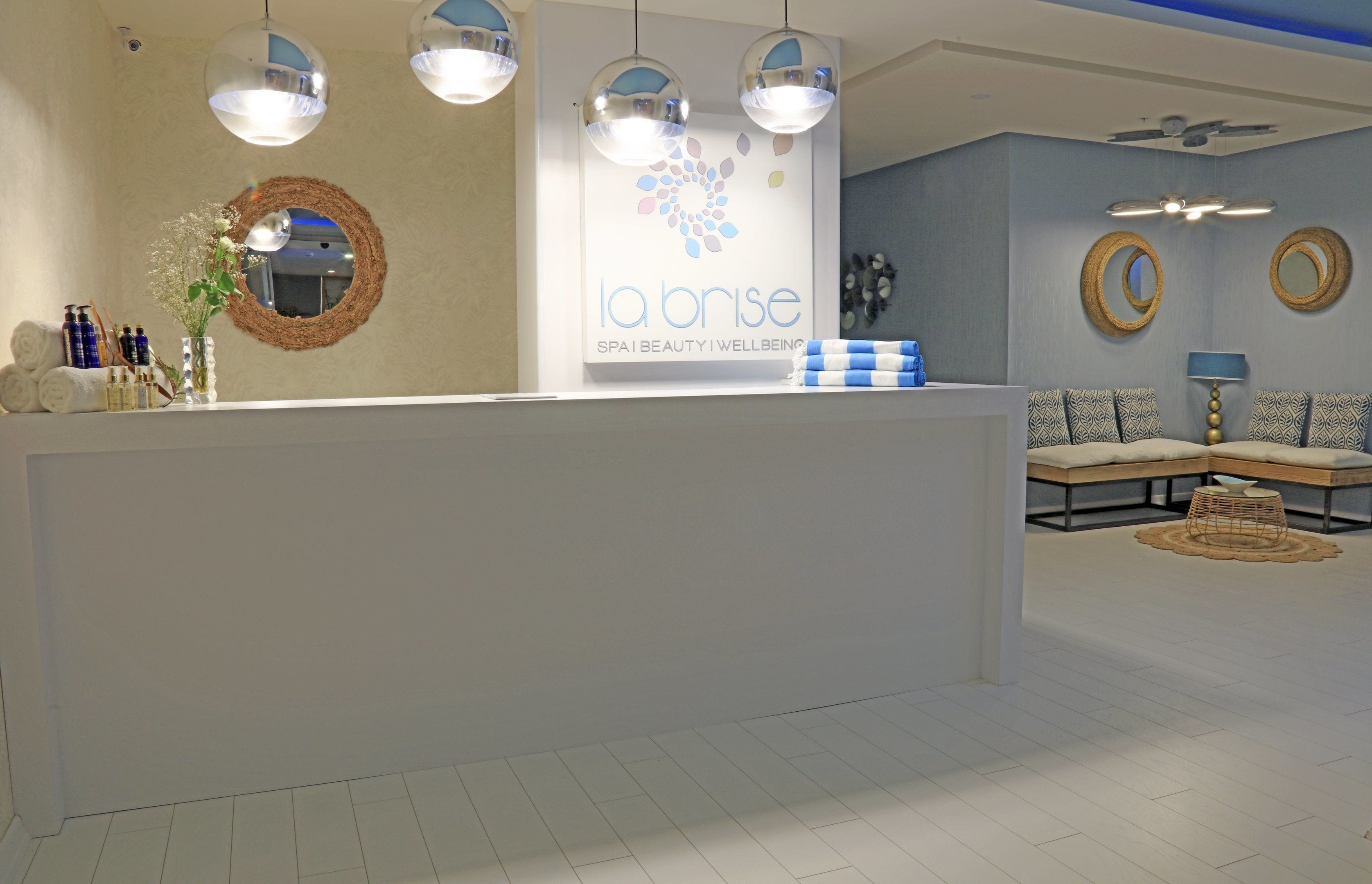 A spa reception area with a white reception desk, hanging pendant lights, and a wall-mounted sign reading "la brise spa beauty wellbeing."