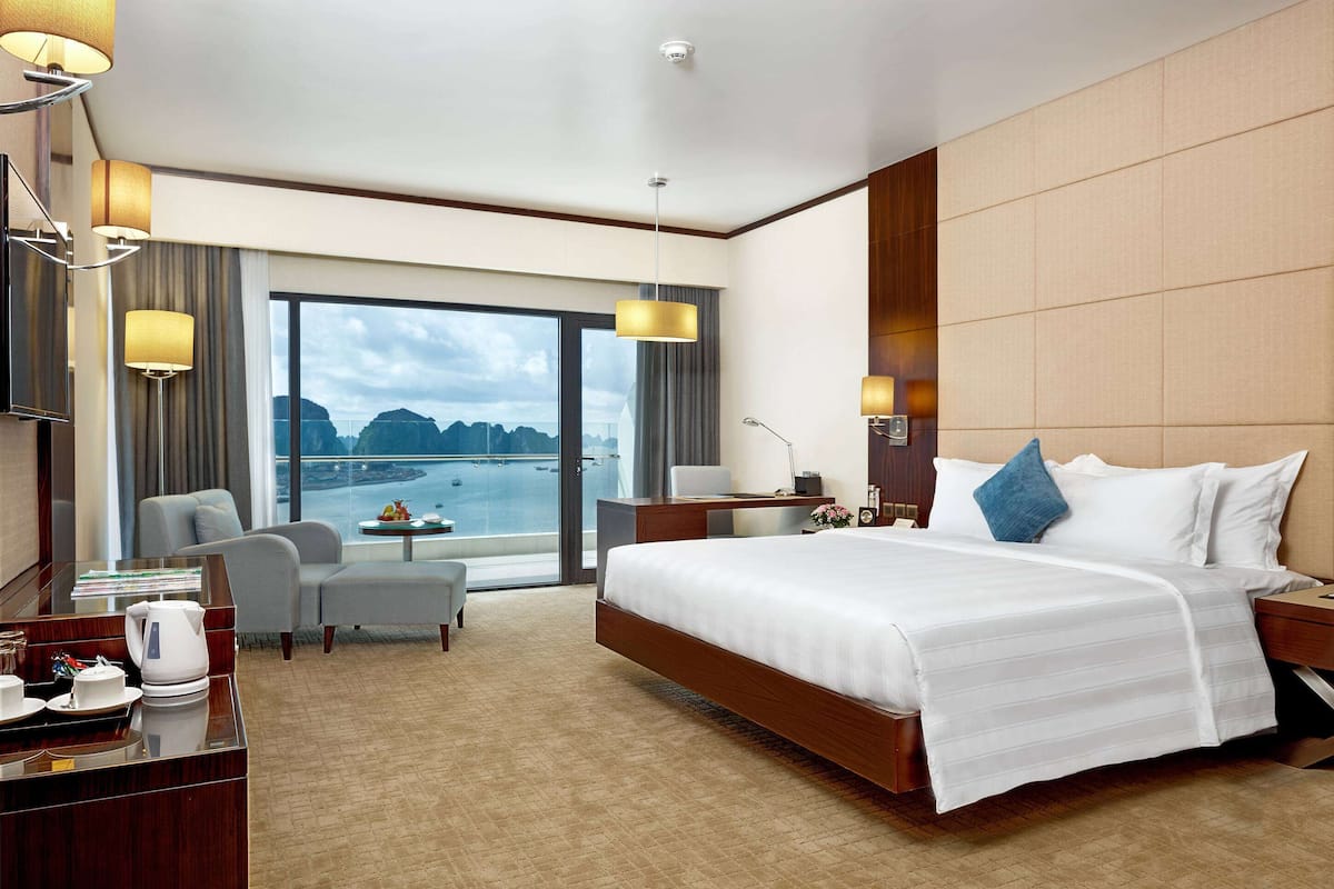 A hotel room with a large bed, a desk with a chair, a small table with a flower vase, and a view of the sea.