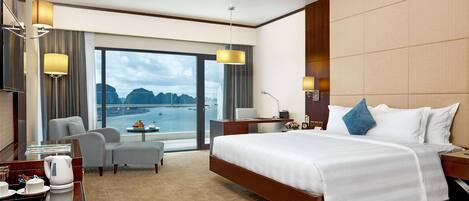 A hotel room with a large bed, a desk with a chair, a small table with a flower vase, and a view of the sea.