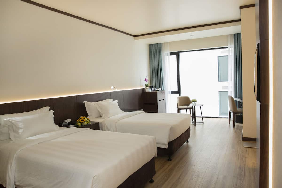 A hotel room with two beds, a small table with a chair, a desk, and a large window with curtains.