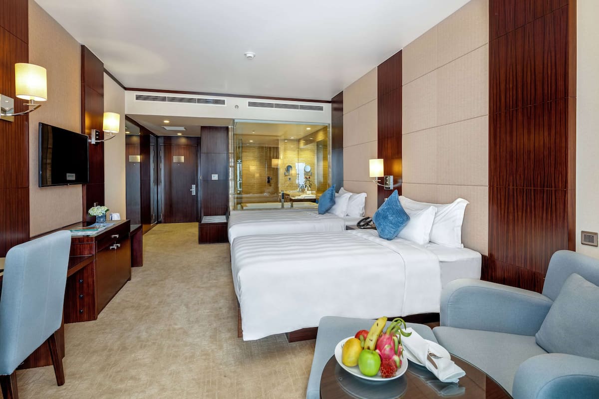 A hotel room with two beds, a desk, a TV, and a dining table with fruit.