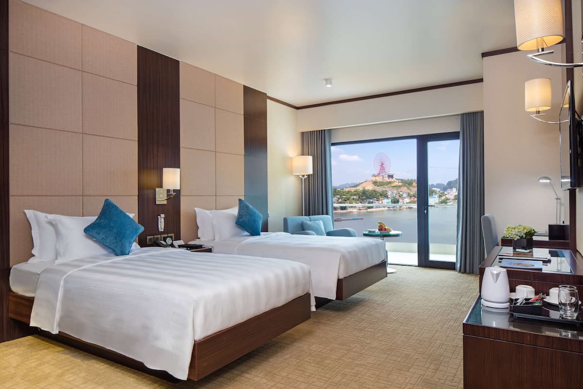 A hotel room with two beds, a desk, a chair, and a large window with a view of a cityscape and a Ferris wheel.