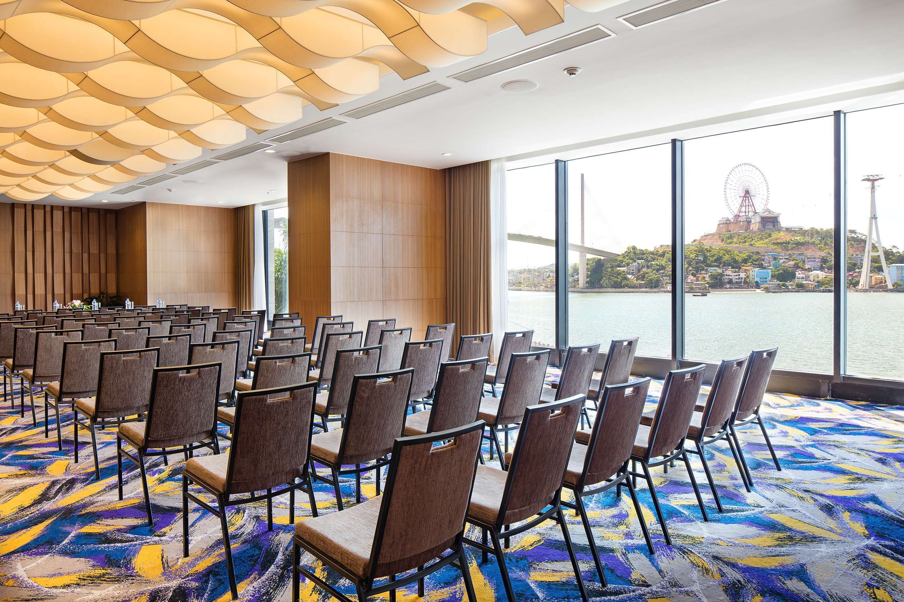 A conference room with rows of chairs and a large window overlooking a body of water with a bridge and Ferris wheel.