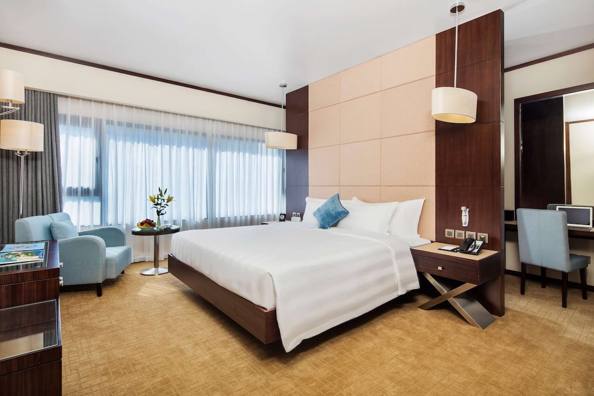 A hotel room with a large bed, a desk with a computer, a chair, a side table with a lamp, and a view of the city through the window.