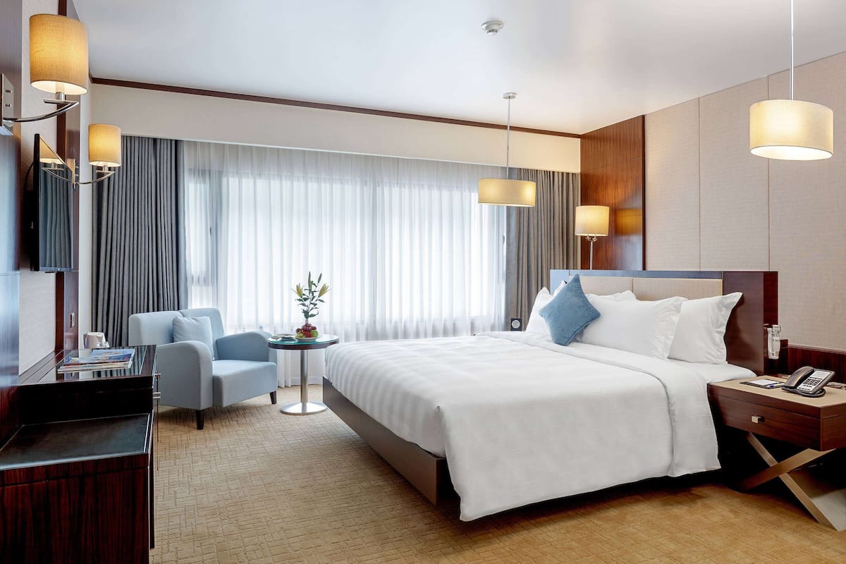 A hotel room with a bed, a desk, a chair, a small table with a plant, and a nightstand with a phone.