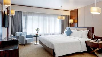 A hotel room with a bed, a desk, a chair, a small table with a plant, and a nightstand with a phone.