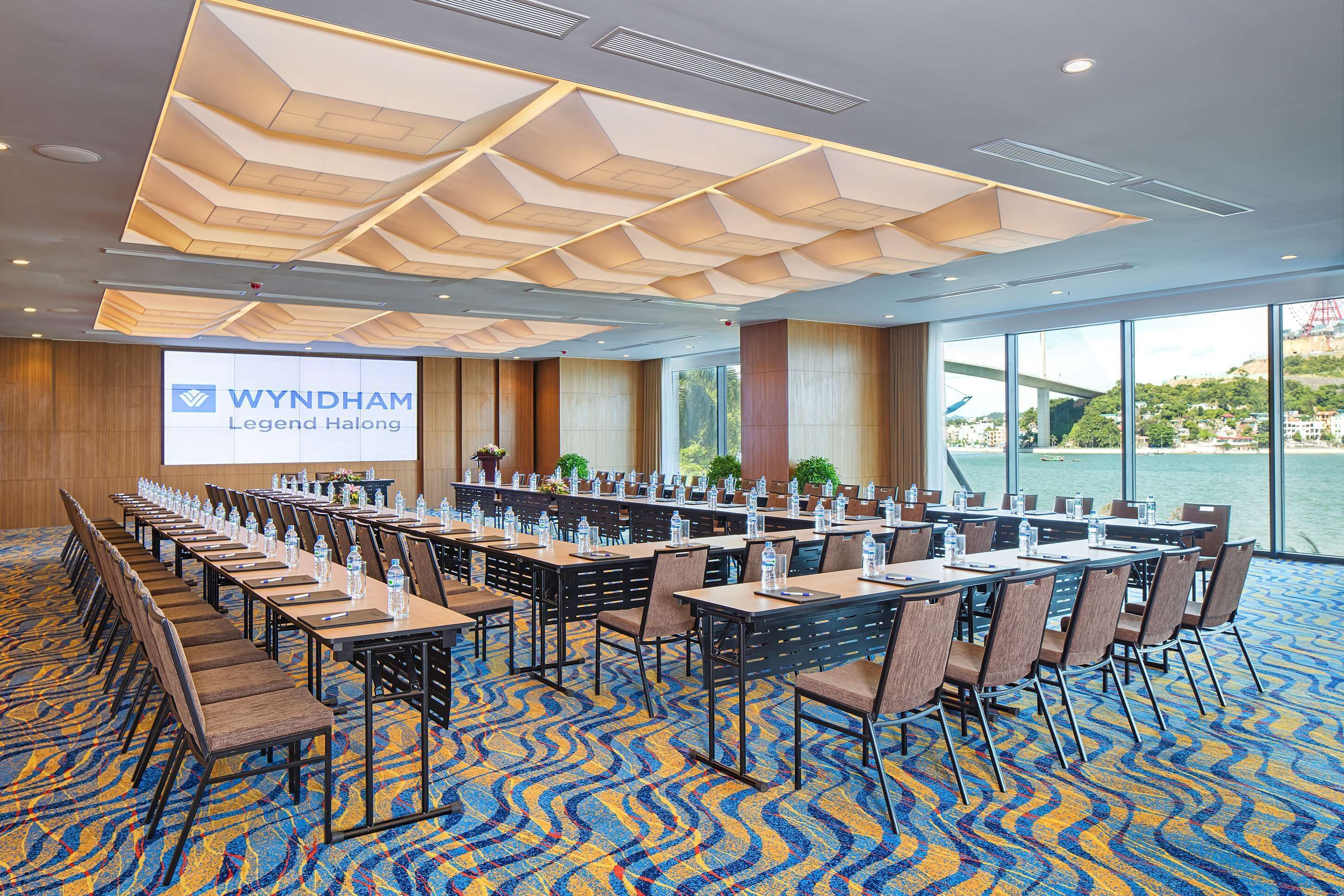 A conference room with a large screen displaying 'WYNDHAM Legend Halong', tables with chairs, and a view of a body of water through large windows.