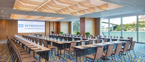 A conference room with a large screen displaying 'WYNDHAM Legend Halong', tables with chairs, and a view of a body of water through large windows.