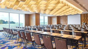 A conference room with rows of tables and chairs, large windows with a view of the sea, and a modern ceiling design.