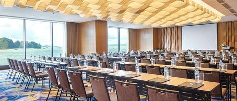 A conference room with rows of tables and chairs, large windows with a view of the sea, and a modern ceiling design.