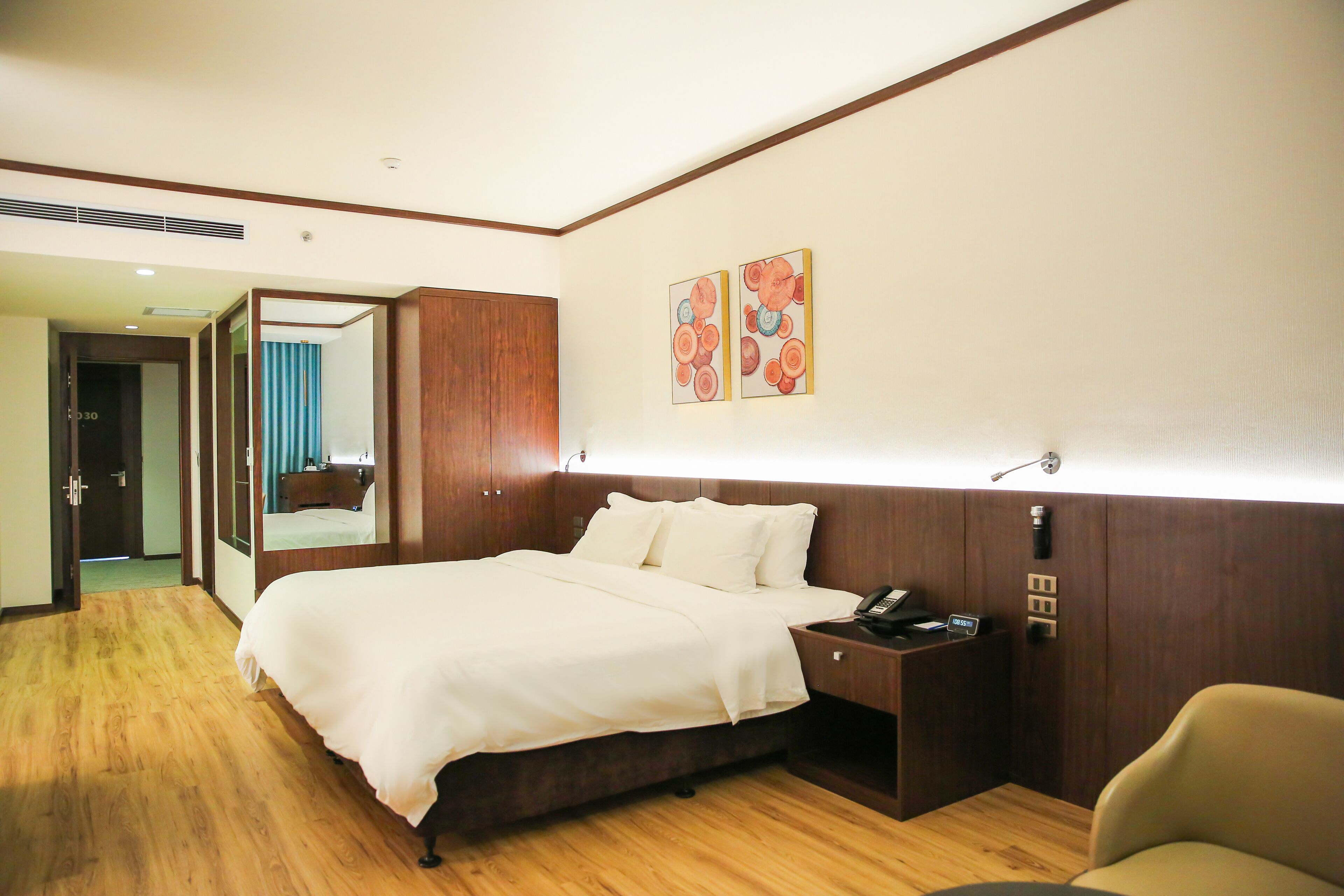 A hotel room with a wooden floor, a large bed with white bedding, a bedside table with a phone, and a mirror.