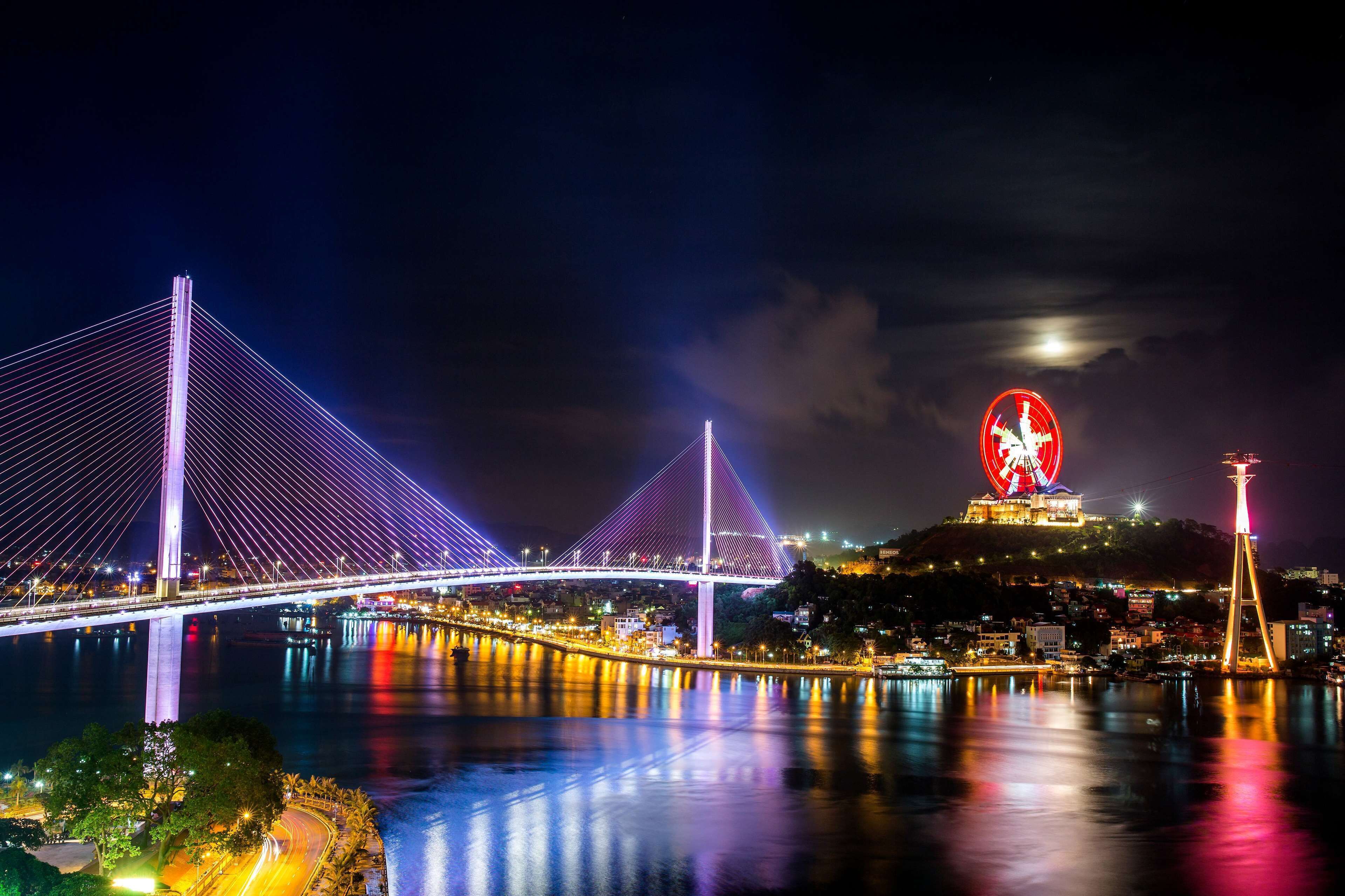 A nighttime view of a bridge with illuminated cables, a lit-up circular structure on a hill, and a body of water reflecting lights.