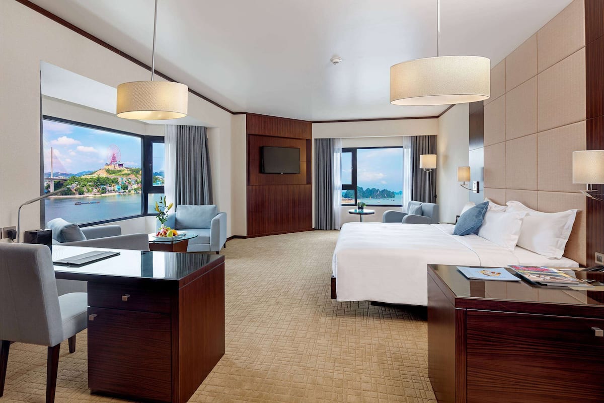 A hotel room with a large bed, a desk, a chair, and a view of a cityscape with a Ferris wheel.