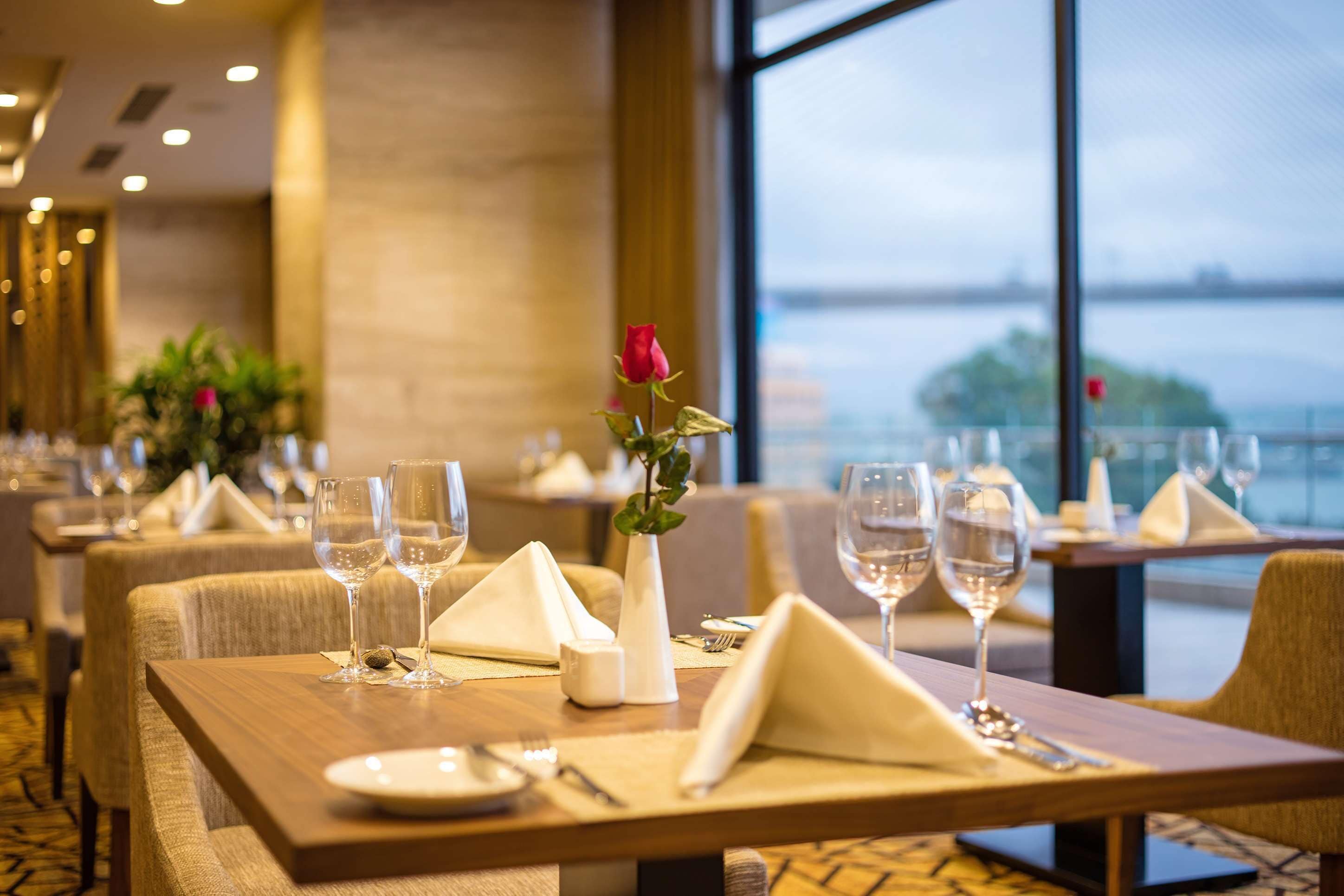 A restaurant with tables set for dining, featuring a vase with a single rose and a view of the water outside.