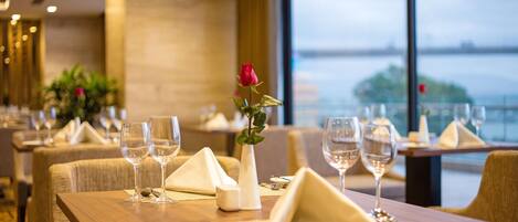 A restaurant with tables set for dining, featuring a vase with a single rose and a view of the water outside.