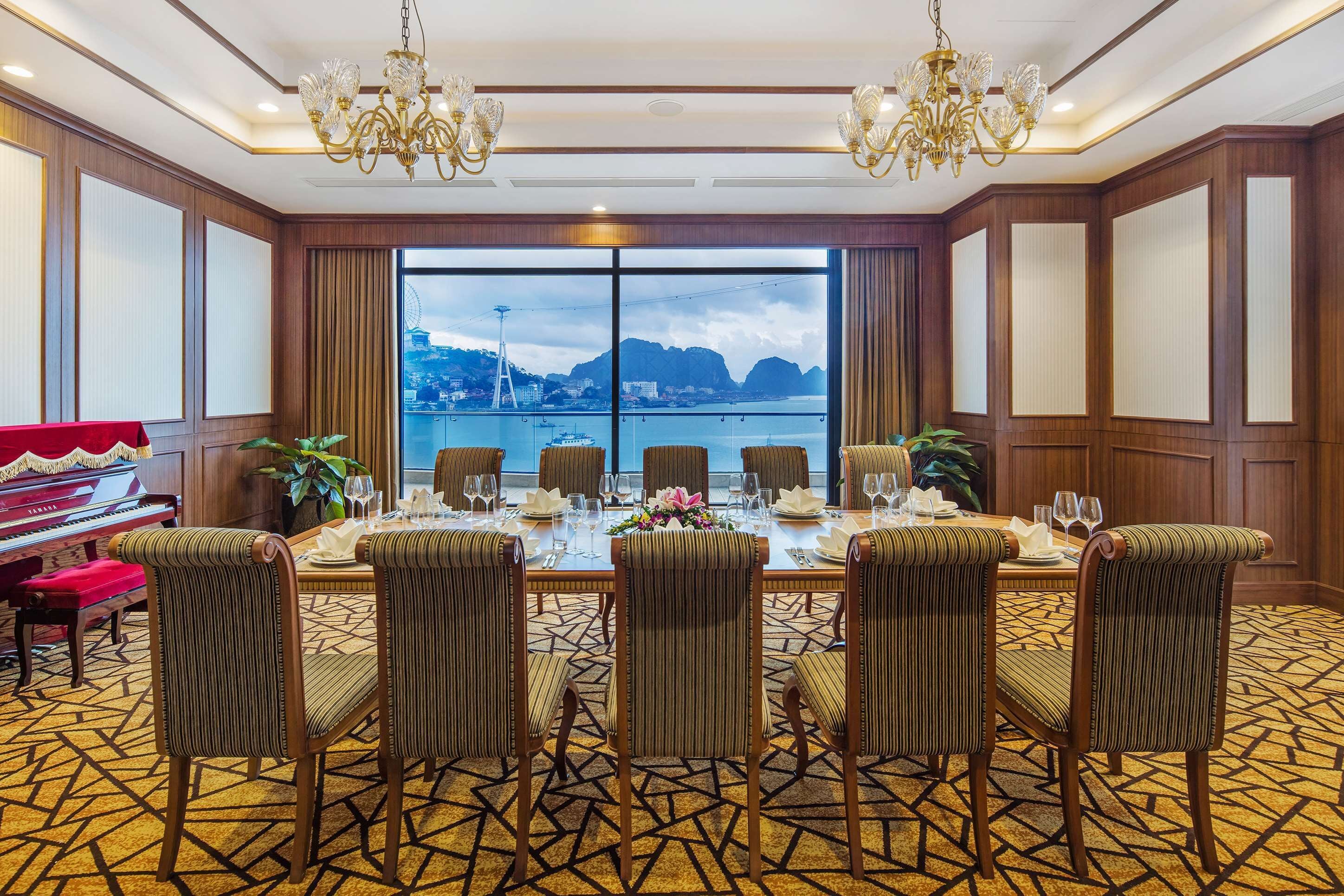 A dining area with a large table set for six, surrounded by six upholstered chairs, with a piano to the left and a view of a body of water and mountains through large windows.