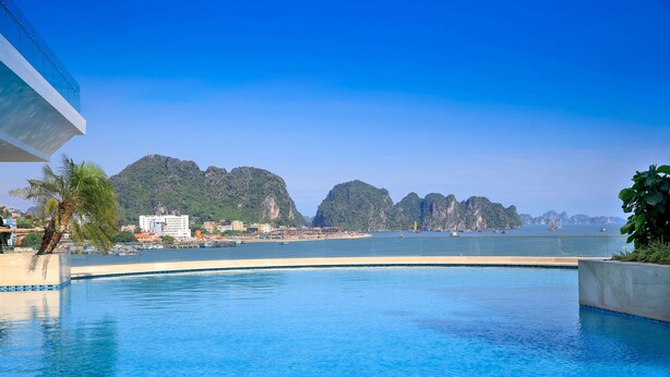 A swimming pool with clear blue water, surrounded by a concrete deck. In the background, there are buildings and a mountainous landscape. - Wyndham Legend Halong Hotel (Bai Chay)
