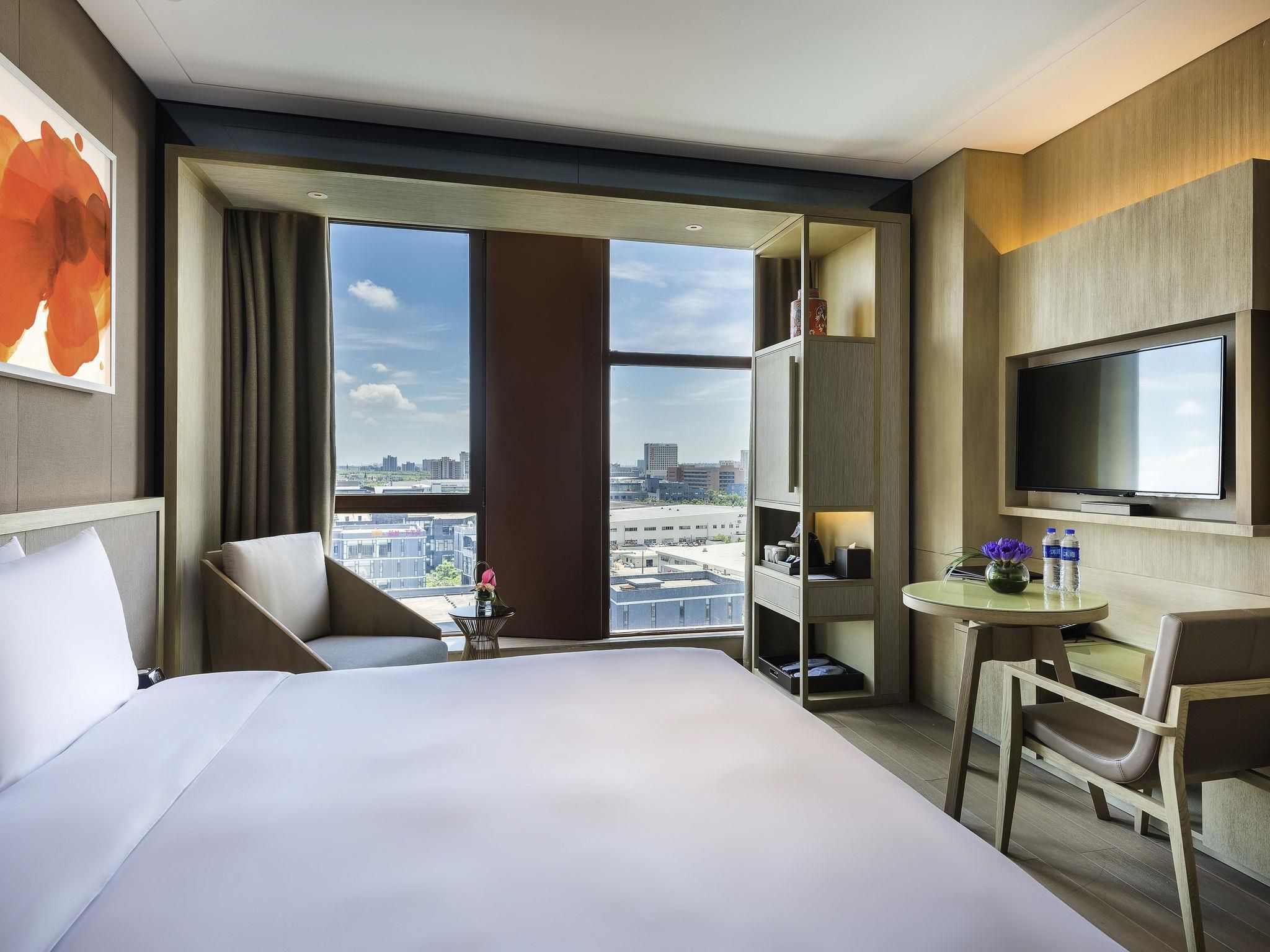 Habitación de hotel con una cama grande, un escritorio con silla, televisión y vista a la ciudad a través de ventanas amplias.