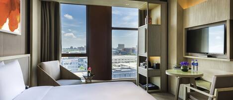 Habitación de hotel con una cama grande, un escritorio con silla, televisión y vista a la ciudad a través de ventanas amplias.