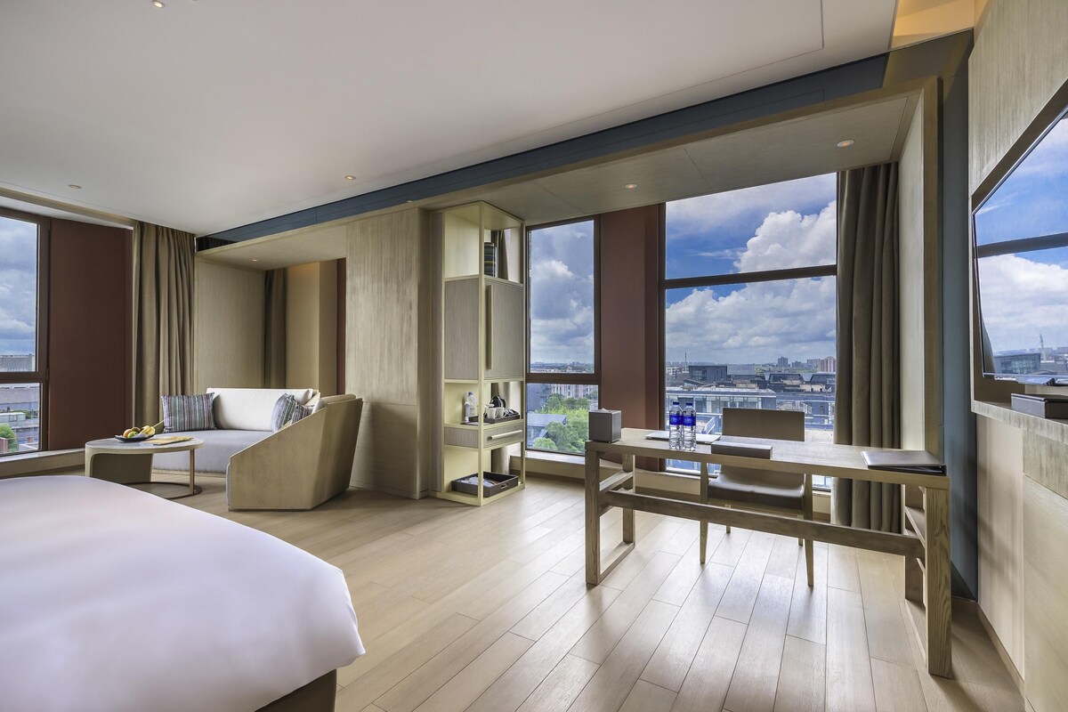 Ein modernes Hotelzimmer mit einem großen Bett, einer Sitzecke, einem Schreibtisch und Blick auf die Stadtlandschaft.