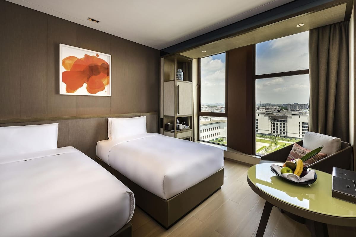 A hotel room with two beds, a desk with a laptop, a fruit bowl, and a large window with a city view.