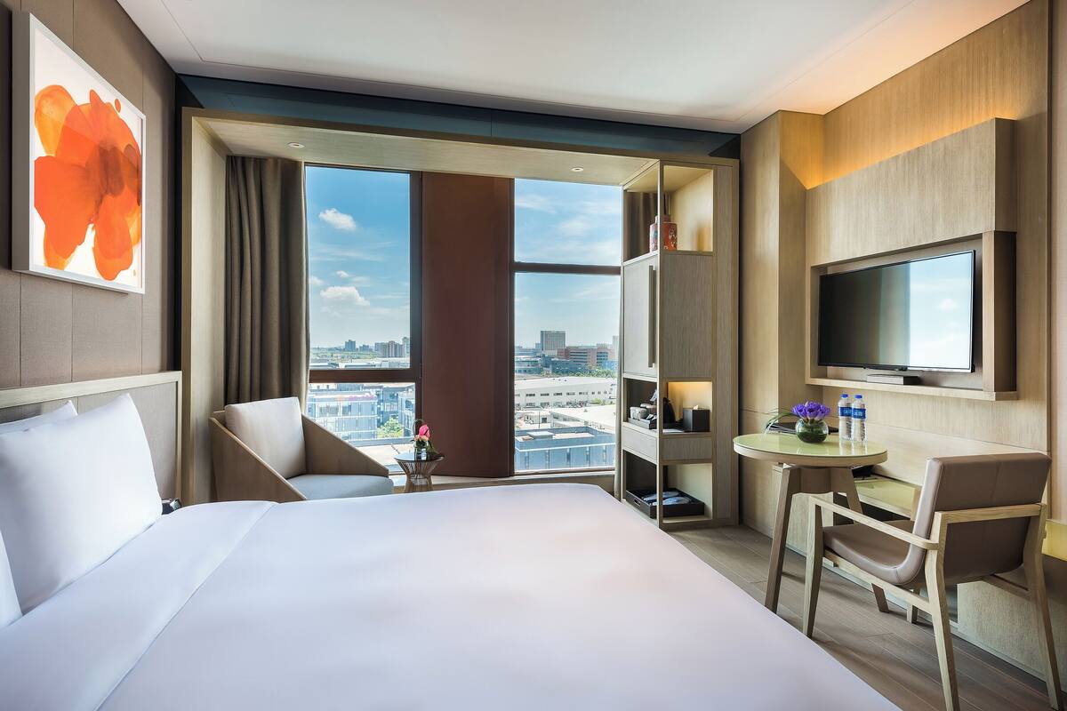 A hotel room with a bed, a desk, a chair, a TV, and a large window with a city view.