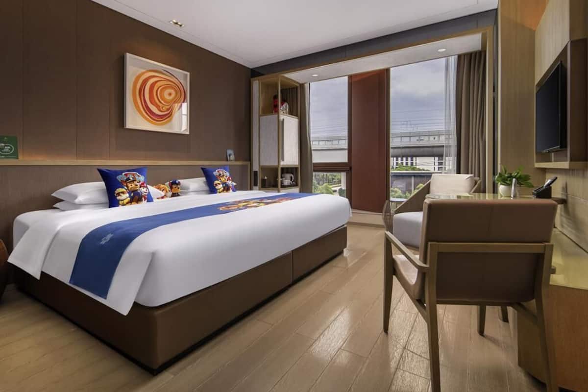 A hotel room with a large bed, a desk, a chair, a TV, and a window with a view of a building.