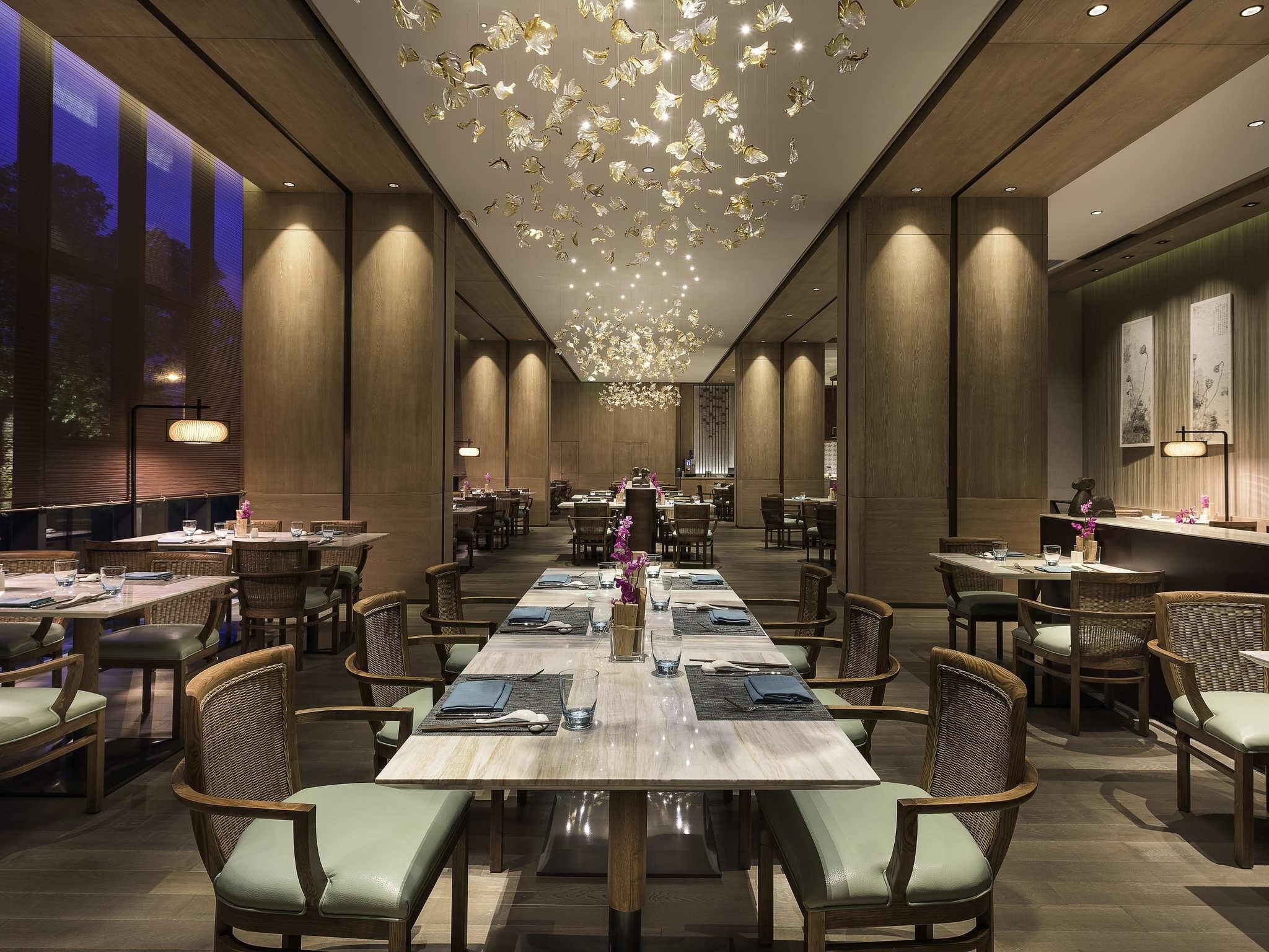 A spacious dining area with long tables and wicker chairs, featuring a modern chandelier and wooden paneling.