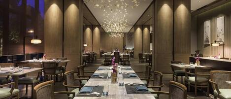 A spacious dining area with long tables and wicker chairs, featuring a modern chandelier and wooden paneling.