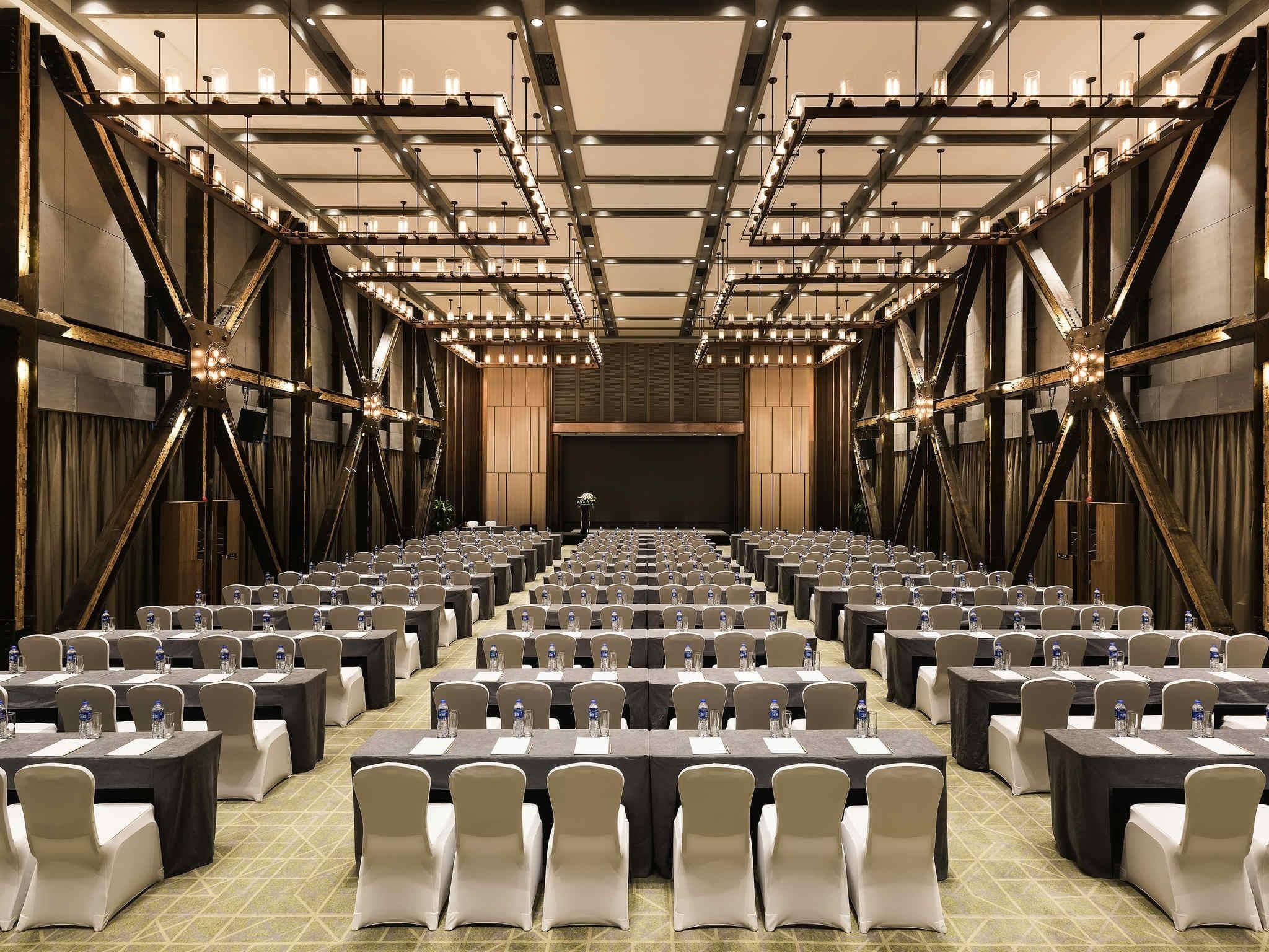 A conference room with rows of tables and chairs, a stage at the front, and a modern industrial ceiling design.