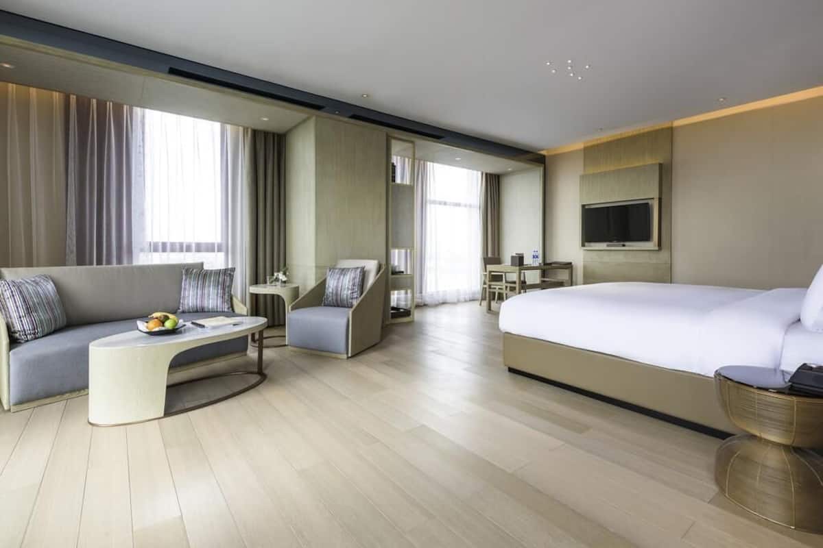 A modern hotel room with a large bed, a sofa, a curved coffee table, a desk, and a TV.