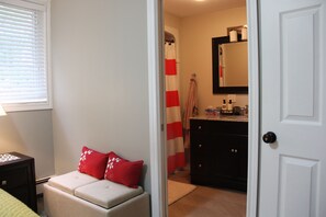 A bedroom with a bed, a nightstand, a desk, and a chair. There is a window with blinds, a door, and a bathroom visible through an open door.