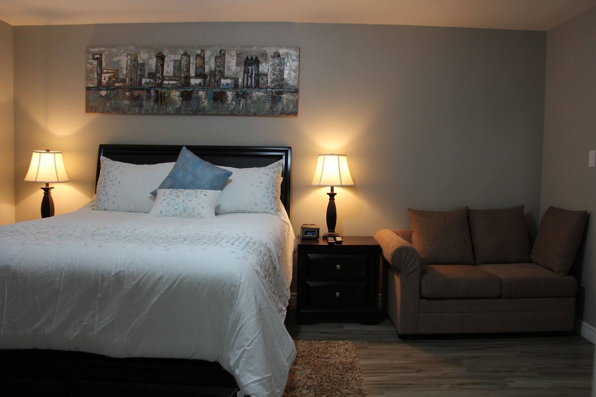 A bedroom with a bed, two bedside tables with lamps, a sofa, and a cityscape painting on the wall.