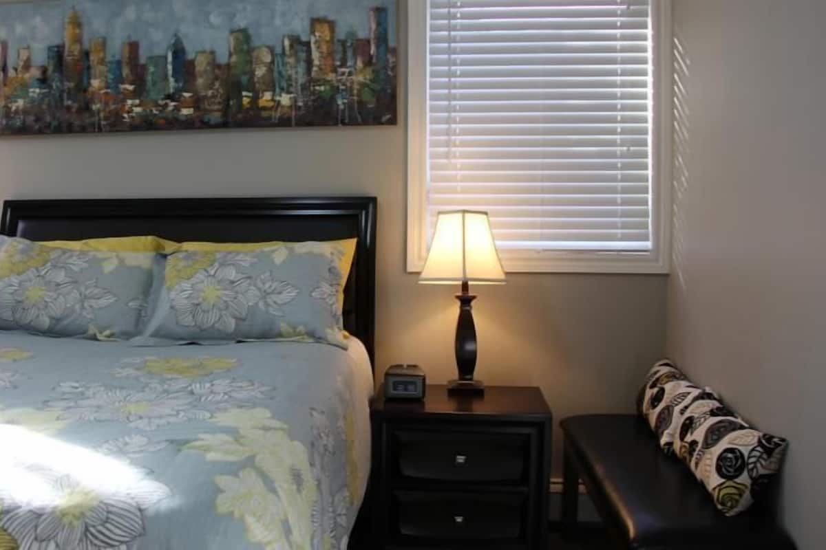 A neatly arranged bedroom with a bed, two bedside tables with lamps, a bench, a cityscape painting, and a window with blinds.