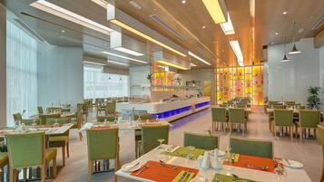 A modern dining area with multiple tables and chairs, a central counter, and a brightly lit wall with shelves.