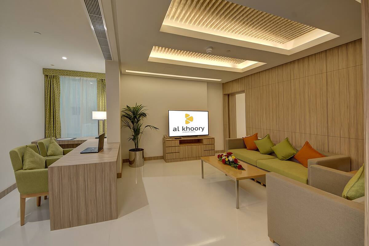 A modern hotel room with a reception desk, a green sofa set, a wooden coffee table, and a flat-screen TV displaying the hotel name.