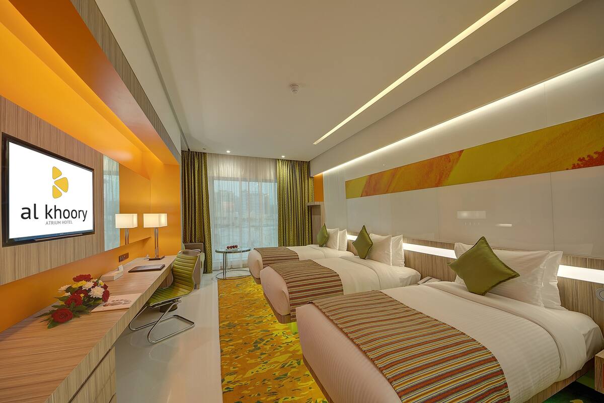 A hotel room with a reception desk, two beds with striped bedding, a desk lamp, a small table with a vase of flowers, and a large window with curtains.