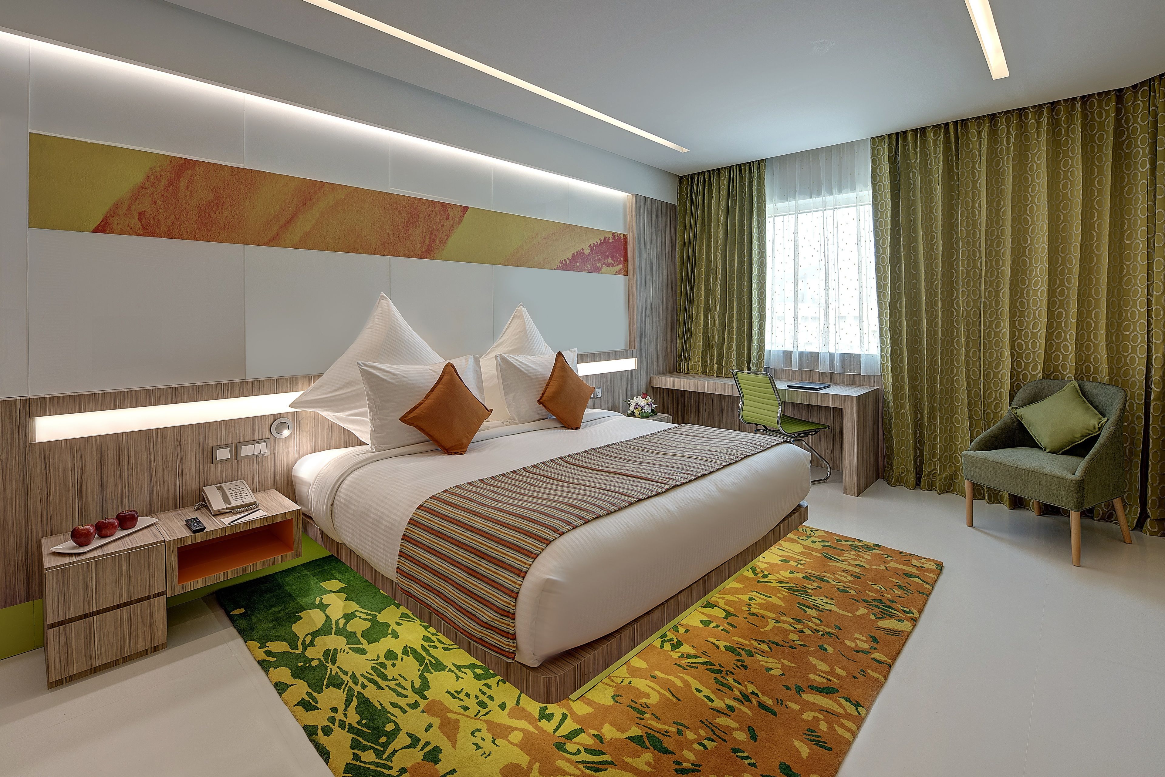 A modern hotel room with a large bed, bedside tables, a desk, and a green armchair.